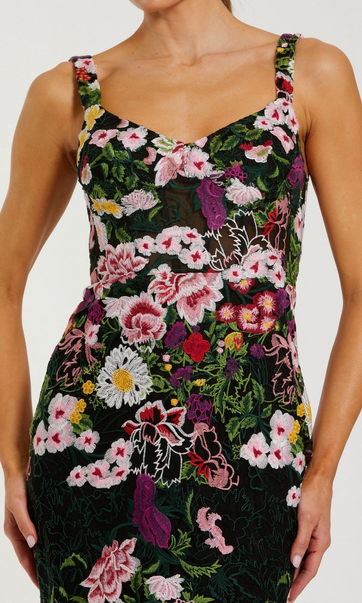 A woman wearing the Mac Duggal 69004 long formal dress, a fitted black gown embellished with vibrant floral embroidery in shades of pink, purple, yellow, and white, along with green leaves. The dress has a subtle mermaid silhouette that accentuates her figure. Her arms are at her sides.